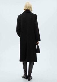 Cappotto di lana nera con un design strutturato, maniche lunghe e colletto a punta. Abbinato a stivali neri in pelle al ginocchio e una piccola borsa.