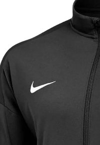 Schwarze Zip-Jacke aus glattem Material, mit einem weißen Nike-Logo auf der linken Brust und einem hohen Kragen für zusätzlichen Schutz.