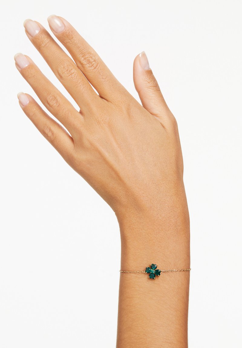 Bracciale in catena d'oro con un mazzo di gemme verdi a forma floreale. Mano con unghie naturali che mostra il gioiello su uno sfondo semplice.