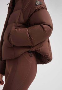 Brązowa kurtka puchowa z pikowanym wzorem i elastycznym dołem, zestawiona z ciemnobrązowymi legginsami o wysokim stanie z kieszeniami bocznymi.