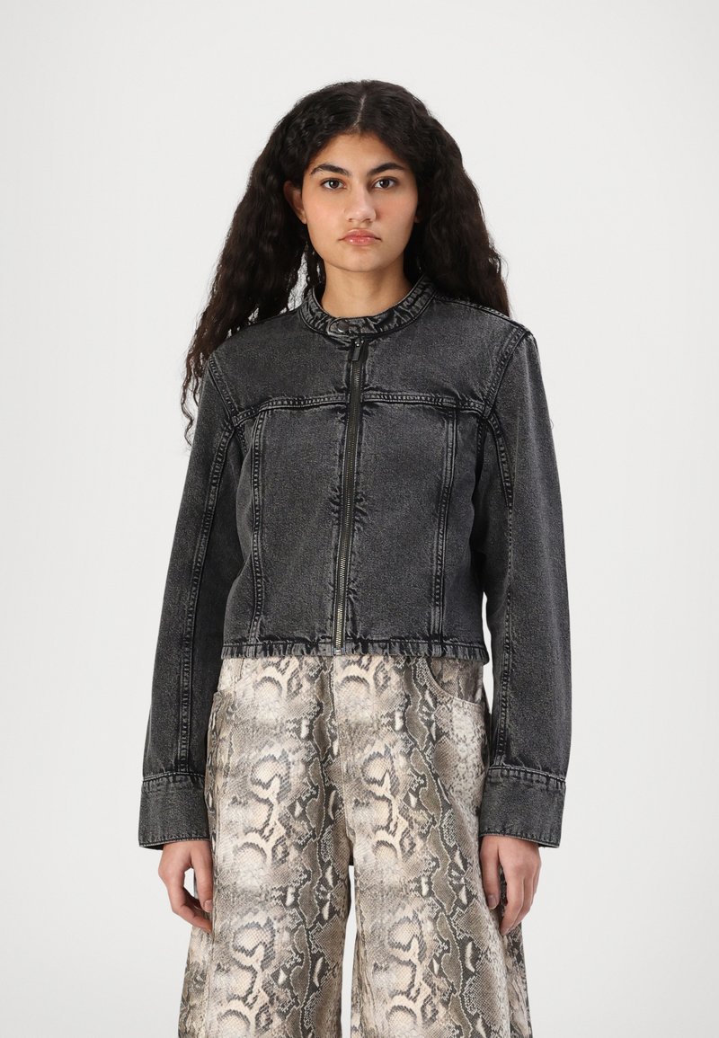 Chaqueta de mezclilla negra recortada con un cuello alto, cremallera frontal y detalles de costura visibles. Combinada con pantalones de pierna ancha con estampado de serpiente.