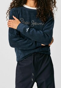 Navy fleece sweatshirt featuring "Pepe Jeans" logo in silver, round neckline, and ribbed cuffs, paired with pinstriped pants.