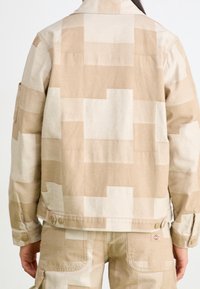 Beige bunda vyrobená z bavlny s patchworkovým vzorem v různých odstínech, s knoflíkovými manžetami a štítkem s logem poblíž lemu.