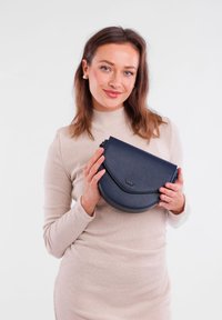 Námořnická kožená crossbody taška s zakřiveným designem, zipovým uzávěrem a embosovaným logem. Měkká textura a minimalistická estetika.