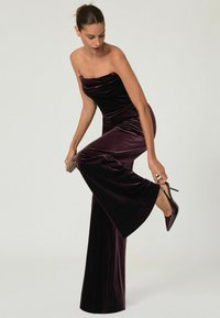 Jumpsuit en velours sans bretelles de couleur violet foncé. Il présente un décolleté drapé et des jambes évasées. Complété par des escarpins à bout pointu et une pochette.