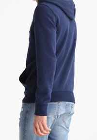 Sudadera azul marino con un bolsillo tipo canguro y puños acanalados. Tejido suave, corte relajado y capucha texturizada. Combinada con vaqueros desgastados de azul claro.