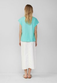 Vrouw met lang blond haar die een turquoise top met korte mouwen, een witte midi-jurk en witte sandalen draagt, staand tegen een effen grijze achtergrond.