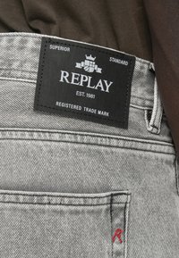 Γκρι τζιν από denim με μαύρη ετικέτα "REPLAY" στη ζώνη μέσης με λευκό κείμενο. Περιλαμβάνει κόκκινο κεντητό λογότυπο στην πίσω τσέπη.