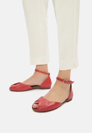 Rote Ledersandalen mit offenem Zehenbereich, überkreuztem Vorderteil und Knöchelriemen. Flache Sohle und glatte Textur, präsentiert mit beigen Hosen.