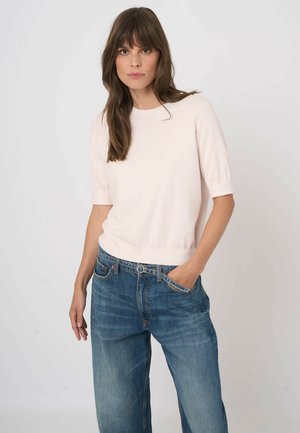 Kobieta o długich brązowych włosach, ubrana w jasnoróżowy krótki top z krótkim rękawem i niebieskie dżinsy, z jedną ręką w kieszeni, na jednolitym tle.