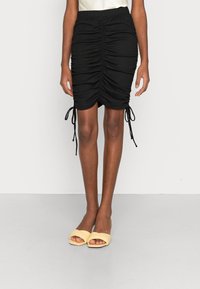 Falda negra con fruncidos y cintas ajustables a los lados, hecha de tela texturizada, combinada con sandalias amarillas de tipo slip-on.