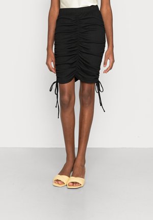 Falda negra con fruncidos y cintas ajustables a los lados, hecha de tela texturizada, combinada con sandalias amarillas de tipo slip-on.