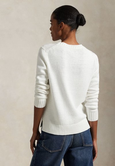 Ženska s temno kožo in lase v figi, oblečena v belo pleteno pulover in modre kavbojke, obrnjena stran na nevtralni podlagi.