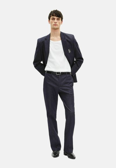 Marineblauer Anzug mit schlanker Passform, strukturiertem Stoff, Ein-Knopf-Blazer und passenden Hosen. Getragen über einem weißen Rundhals-T-Shirt, kombiniert mit schwarzen Schuhen.