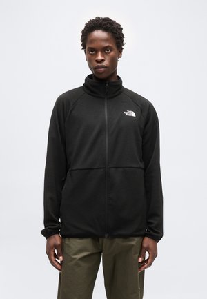 Moški, oblečen v črno flis jakno z zadrgo in logotipom The North Face na prsih ter olivno zelene hlače, stoji pred enobarvnim ozadjem.