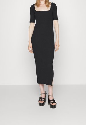 Frau trägt ein schwarzes, geripptes Midikleid mit kurzen Puffärmeln und quadratischem Ausschnitt, kombiniert mit schwarzen Plateausandalen.