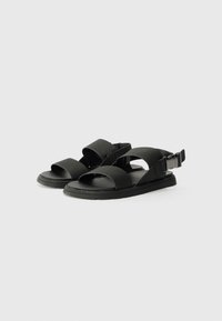 Svarte sandaler med brede stropper og flat såle, laget av mykt materiale, med spenne lukking på baksiden for justerbar passform.
