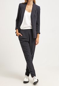 Costume sombre à rayures fines avec un blazer ajusté et un pantalon fuselé. Débardeur blanc en dessous, associé à des chaussures noir et blanc. Texture lisse.