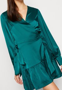 Tyrkysové saténové šaty s dlouhými rukávy, s nařaseným pasem, rozšířeným lemem a bočními zavazováním pro nastavitelný střih. Hladká textura a splývavý design.