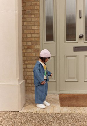 Tout-petit portant une tenue en denim et un bonnet rose se tient en regardant vers le bas près d'une porte beige avec un paillasson devant une maison en briques.