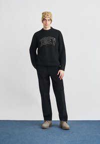 Čierny pletený sveter s logom "OBEY", doplnený čiernymi nohavicami a béžovými čižmami. Modelka má na hlave béžovú čiapku s bodkami. Jednoduché pozadie.