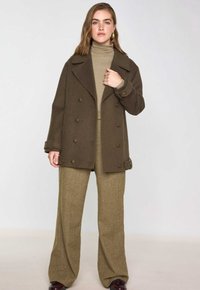 Cappotto doppio petto in lana marrone con grandi bottoni, indossato sopra un dolcevita beige e pantaloni larghi con trama.