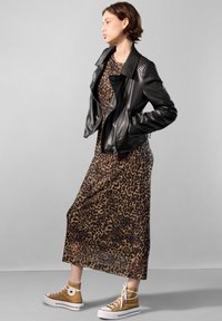 Giacca da motociclista in pelle nera con spalle trapuntate indossata sopra un vestito maxi con stampa leopardata, abbinata a sneakers alte beige.
