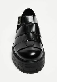 Black leather sandal with woven straps, rounded toe, adjustable buckle, and thick, ridged sole. Smooth texture with a glossy finish.