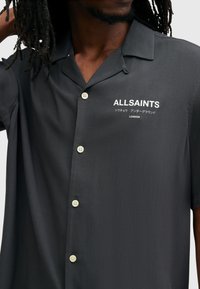 Zwart shirt met korte mouwen van een soepele stof, met een knoopsluiting aan de voorkant en witte logotext op de linkerborst. Subtiele glans.