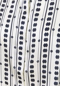 Tissu blanc avec un motif répété de cercles noirs et de lignes, d'une texture lisse et avec une fermeture par bouton au centre.