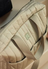 Beige gewatteerde totebag met stoffen handvatten, met een voorzak en een rechthoekige vorm. Metallic logo detail aan de zijkant.
