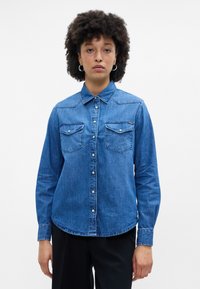 Camisa de ganga abotoada em azul médio, com colarinho, dois bolsos no peito com aba e uma bainha arredondada. Textura suave e design clássico.