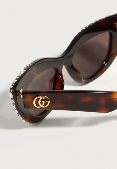 Γυαλιά ηλίου με σκελετό σε σχέδιο χελωνί και φακούς με σκούρο χρώμα, διακοσμημένα με στρας στο πάνω μέρος της κορνίζας και χρυσλό λογότυπο διπλού G στο μπράτσο.