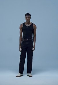 Top preto sem mangas, jeans escuros, ténis brancos. Modelo tem cabelo curto e usa um colar de corrente prateada em frente a um fundo azul claro.