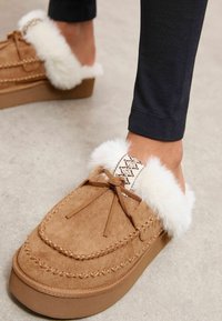 Zapatilla de ante marrón con ribete de piel sintética blanca y costura decorativa. Presenta un lazo y una suela marrón acolchada.