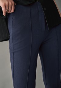 Pantaloni eleganți bleu marin, cu o textură fină, având un buzunar lateral și detalii cu cusături verticale. Purtați cu un sacou negru cu dungi.