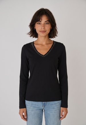 Donna con capelli corti e scuri che indossa una maglietta nera a maniche lunghe con scollo a V e piccoli borchietti, e jeans azzurri chiari, in piedi davanti a uno sfondo semplice.