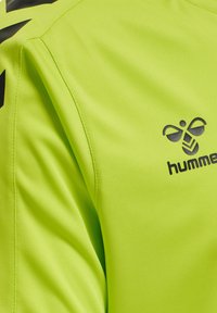Limegrøn atletisk skjorte lavet af glat stof, med et sort logo og et karakteristisk skulderdesign med kontrasterende accenter.