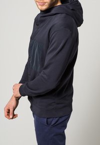 Námořnická modrá mikina s lesklou černou kapsou na prsou, knoflíkovým zapínáním a žebrovanými manžetami. Má uvolněný střih a jemnou texturu látky.