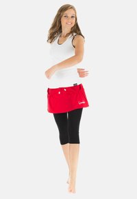 Rote Rock mit Taschen, gebundenem Bund und weißem Logodetail, kombiniert mit einem weißen Tanktop und schwarzen Capri-Leggings, Modell in Bewegung.