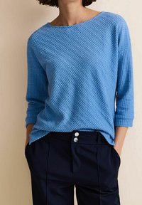Blå strukturerad stickad tröja med båturringning och trekvartslånga ärmar, kombinerad med mörkblå byxor med två knappar vid midjan.