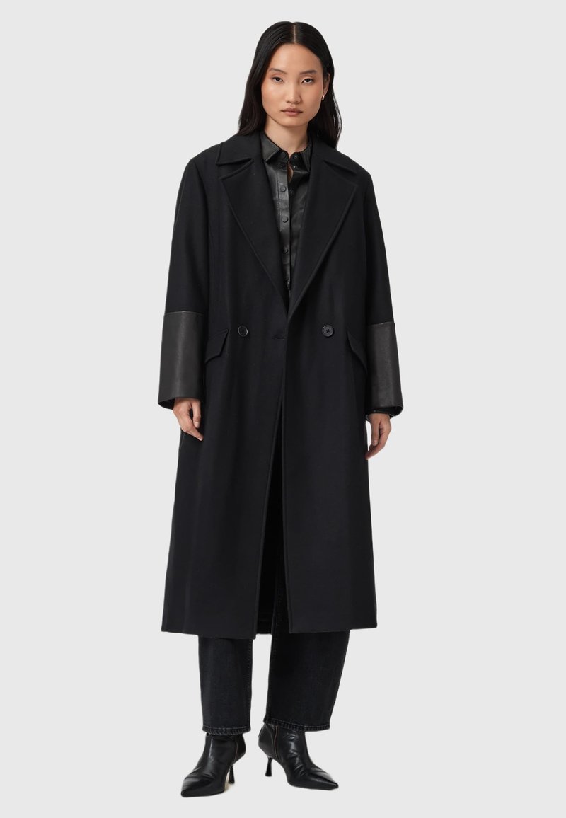 Cappotto lungo nero con ampie rever, bottoni doppiopetto e maniche in finta pelle. Indossato sopra una camicia nera e abbinato a stivaletti alla caviglia.