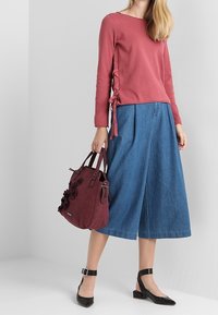 Rotes Langarm-Oberteil mit seitlichem Bindedetail, kombiniert mit einem blauen Denim-Culotte-Rock. Schwarze Ballerinas mit Absatz und eine kastanienbraune Handtasche mit floralen Akzenten.