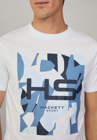 Camiseta blanca de manga corta con un diseño geométrico abstracto en azul y el texto "HS" y "HACKETT SPORT" en azul marino y negro.
