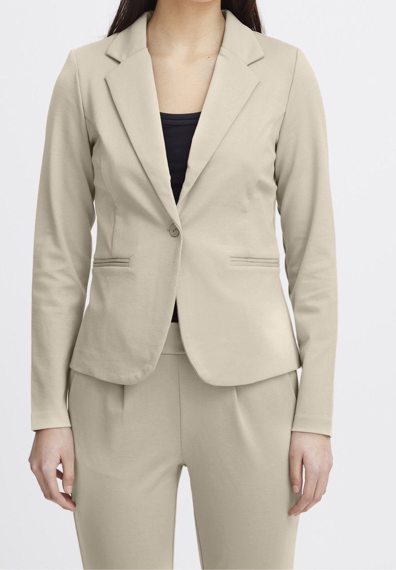 Femme portant un blazer beige ajusté et un pantalon assorti avec un top noir en dessous, debout sur un fond uni.