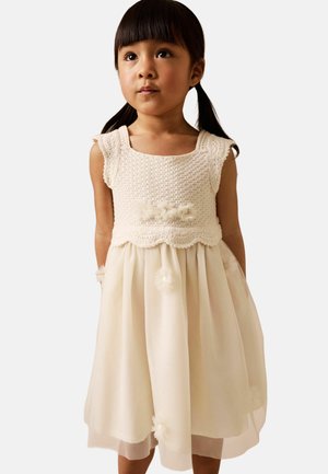 Junges Mädchen mit dunklen Zöpfen, das ein cremefarbenes, ärmelloses Kleid mit gehäkeltem Oberteil und einem Tüllrock trägt, der mit Stoffblumen verziert ist.