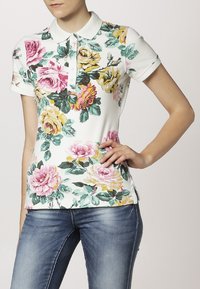 Polo shirt floral en tissu blanc avec des roses roses et jaunes, des feuilles vertes et un col classique. Manches courtes et fentes sur les côtés.