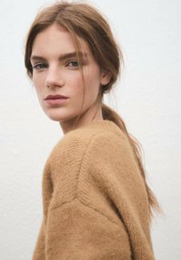 Un suéter de punto de color marrón claro con textura y un corte relajado, que muestra una costura en el hombro y un aspecto suave, usado con el cabello suelto.