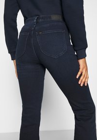 Person klädd i mörkblå slim-fitjeans med bakfickor, stående med en hand vilande på låret framför en neutral bakgrund.