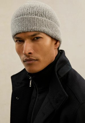 Man wearing a gray knit beanie and a black jacket with a high collar, looking directly ahead against a neutral background.